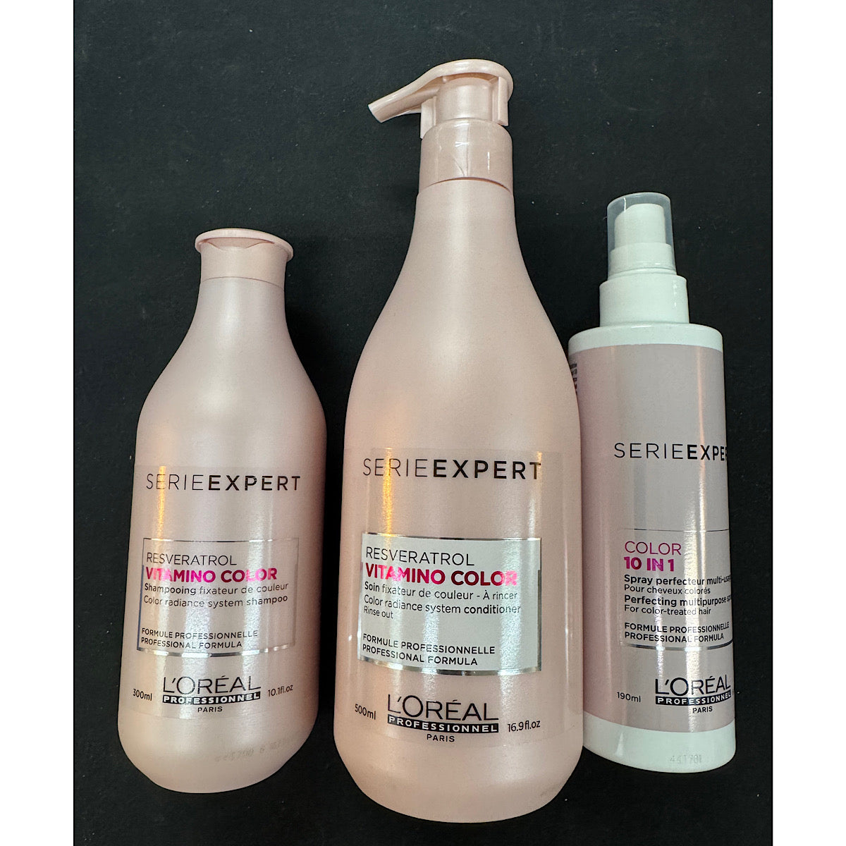 Three L'Oréal Professionnel L'Or Élite Series Expert haircare bottles in a set of three, featuring a Reversatrol Vitmino Color shampoo, conditioner, and spray on a dark background.