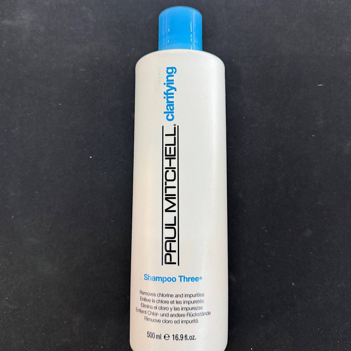 A 500 ml white bottle of Paul Mitchell Clarifying Shampoo Three with a bright blue cap, set against a dark textured background.