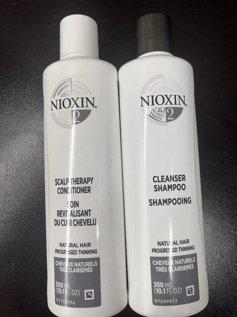 Two white Nioxin hair care bottles on a dark background: Scalp Therapy Conditioner on the left with a white cap and gray label, and Cleanser Shampoo on the right with a black cap and gray label.